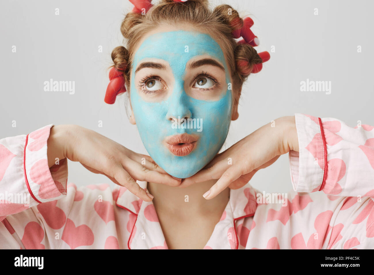 Close-up portrait of european girly girl with cute expression, wearing pyjamas and hair-curlers, standing with facial mask over gray background, holding hands on chin and looking up with folded lips. Stock Photo