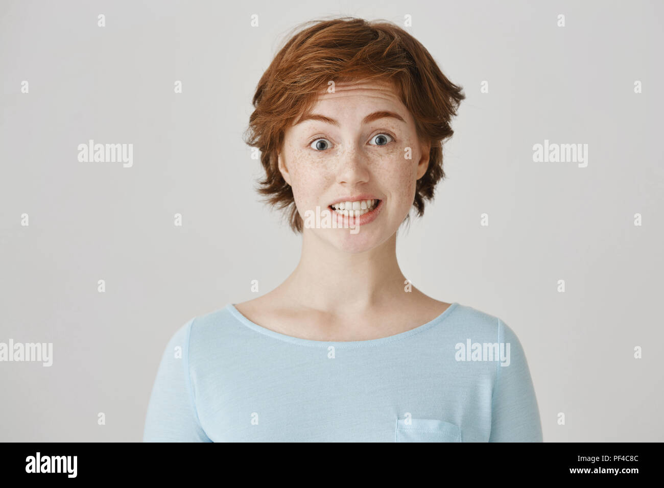 Studio portrait of attractive caucasian female with red hair and freckles feeling awkward, lifting eyebrows and smiling nervously, having no clue what to say in strange situation on meeting Stock Photo
