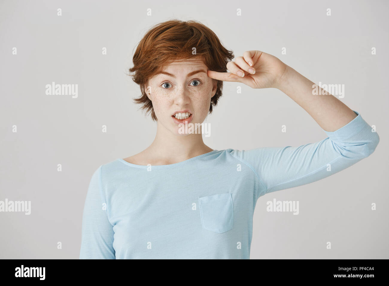 Are you insane or crazy. Portrait of irritated angry woman with ginger hair and freckles rolling index finger near temple, losing temper while seeing friend doing stupid things over gray wall Stock Photo