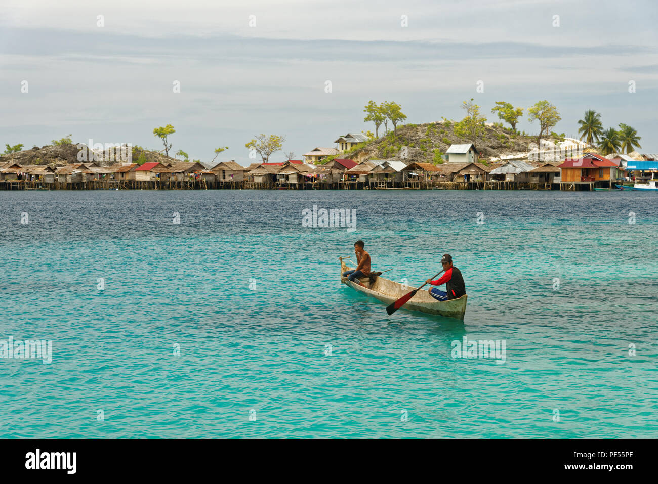 View of two men paddling on a traditional boat in front of  bajo village with wooden houses on the Togean islands in Central Sulawesi, Indonesia Stock Photo