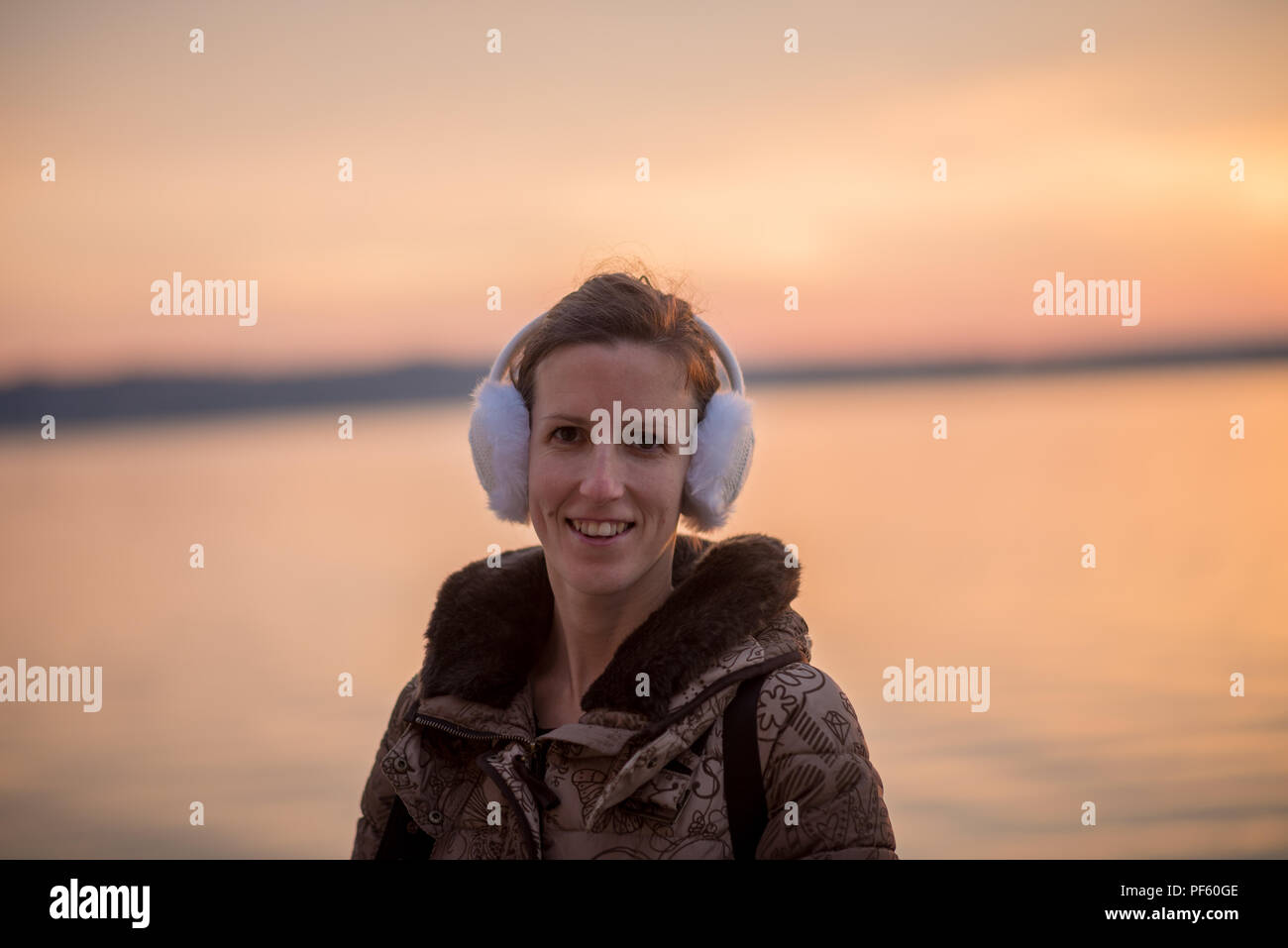 Smiling woman wearing fluffy ear muffs at sunset standing against an ocean as she enjoys a winter evening outdoors. Stock Photo