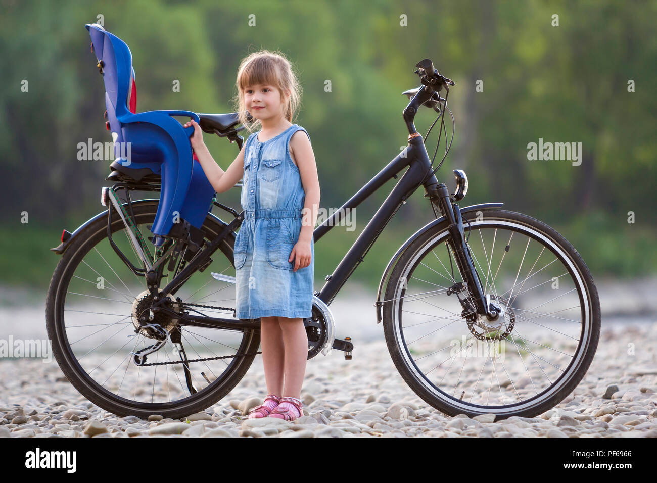 Small pretty blond girl in blue dress standing on pebbles in front of modern bicycle with child seat on blurred green trees background on summer day.  Stock Photo