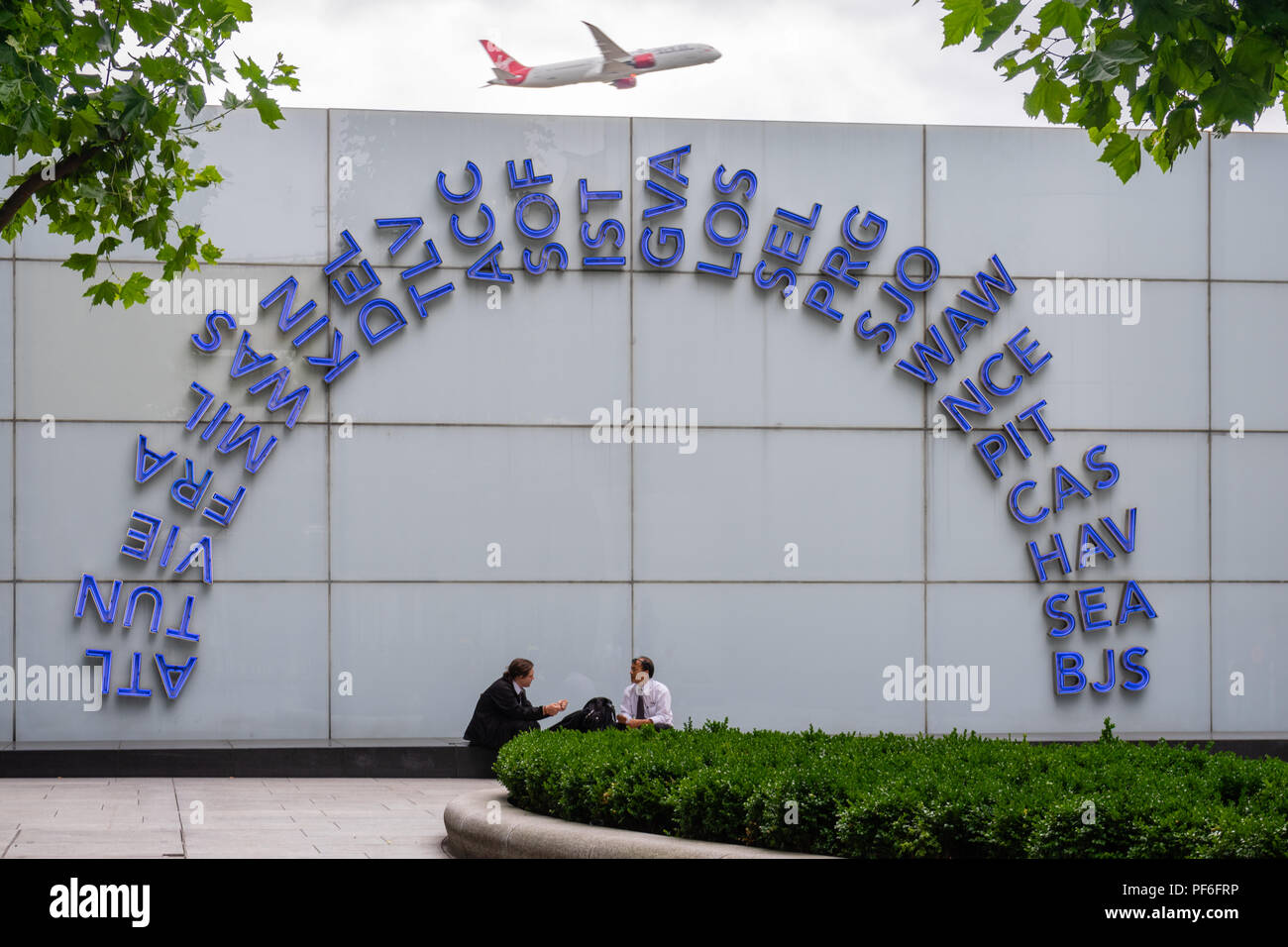 Blue illuminated world IATA codes displayed outside London Heathrow airport terminal 5 building,a Virgin Atlantic plane taking off in the background Stock Photo