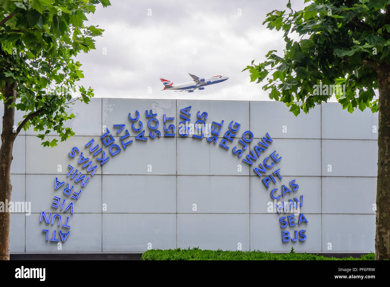 Blue illuminated world IATA codes displayed outside London Heathrow airport terminal 5 building, British airways plane taking off in the background,UK Stock Photo