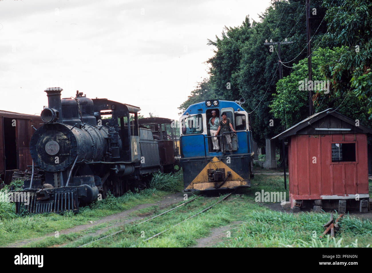 Managua, Nicaragua,, Feb 1990; A railway goods train drives into Managua station, past on old derelict steam engine parked in a siding. Stock Photo