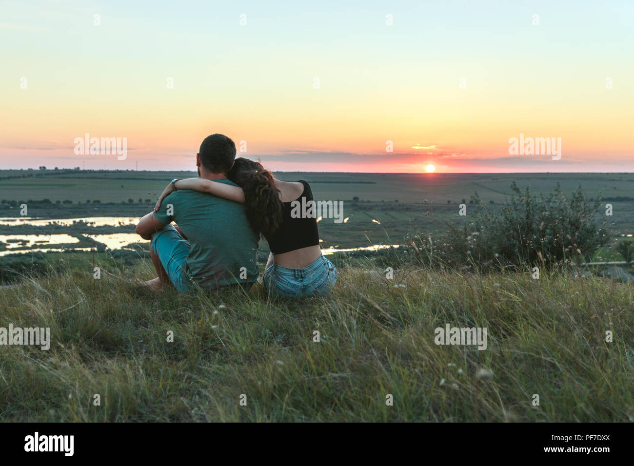 Couple is hugging and sitting close on a hill at sunset Stock Photo