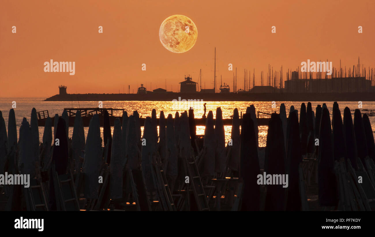 Beautiful full red moon above the harbor seen from the beach with sun umbrellas Stock Photo