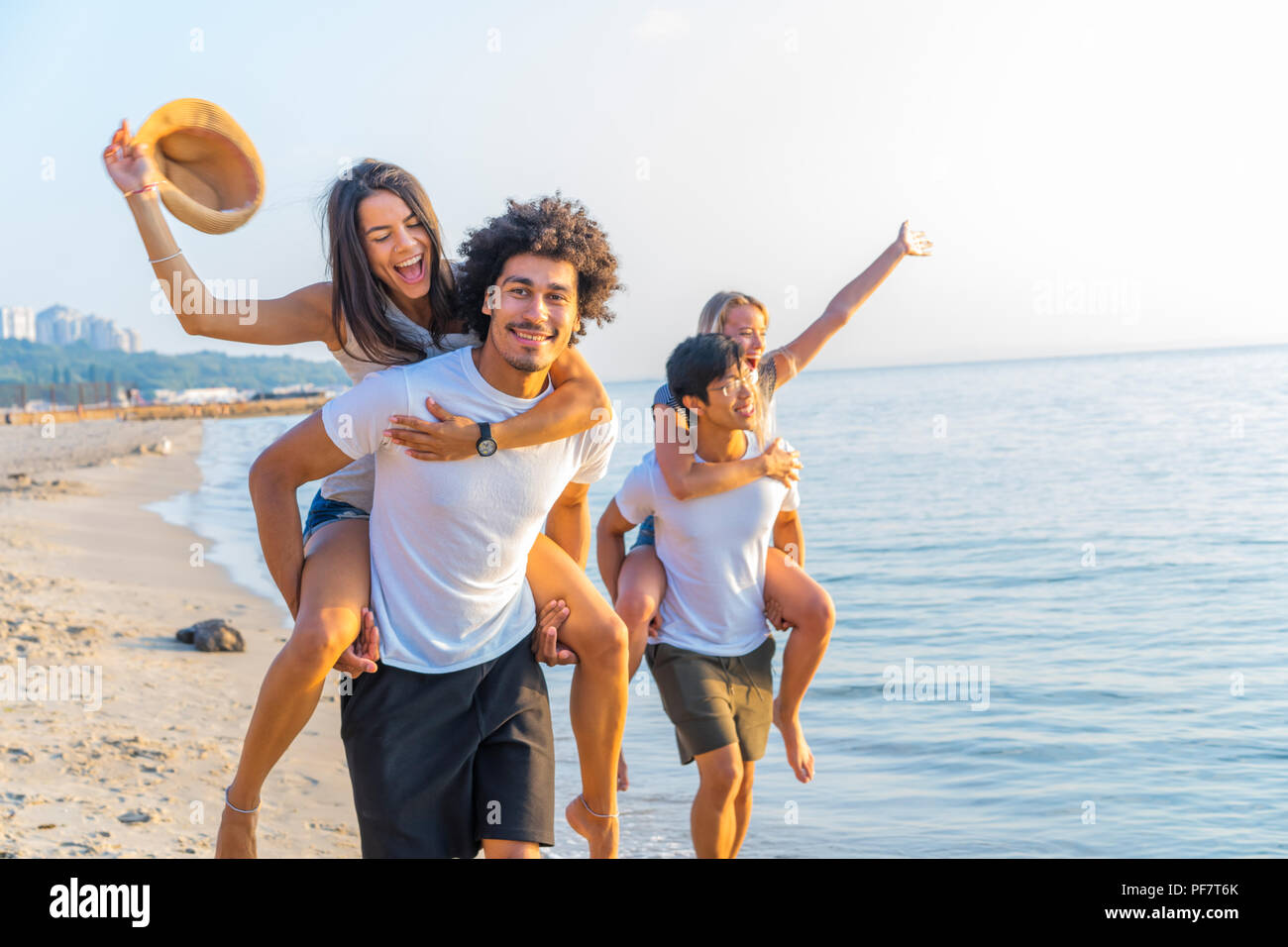 Group of friends walking along the beach, with men giving piggyback ride to girlfriends. Happy young friends enjoying a day at beach Stock Photo