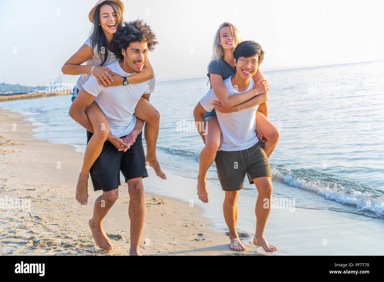 Group of friends walking along the beach, with men giving piggyback ride to girlfriends. Happy young friends enjoying a day at beach Stock Photo