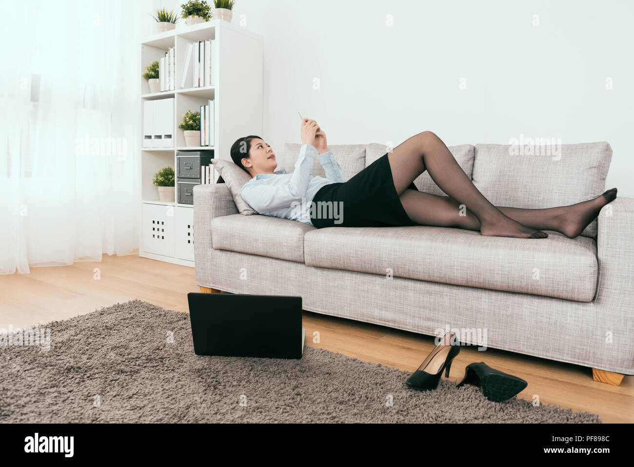 she is playing on her smartphone and also working at home by using notebook online at the same time lying down on cozy sofa Stock Photo