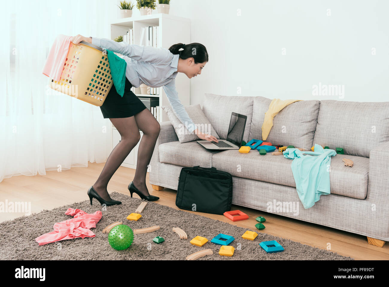 beautiful Japanese businesswoman is using notebook reply the E-mail and cleaning up the mess toys and clothes at home. Stock Photo