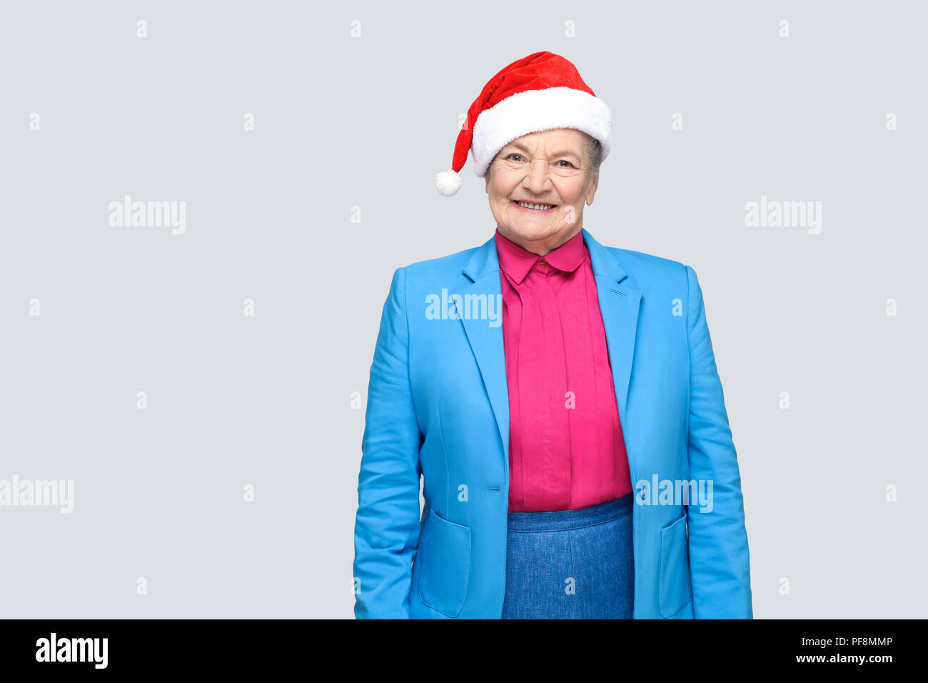 Satisfied toothy smiling colorful casual style aged woman with blue suit and christmas santa red cap standing and looking at camera with joyful face.  Stock Photo