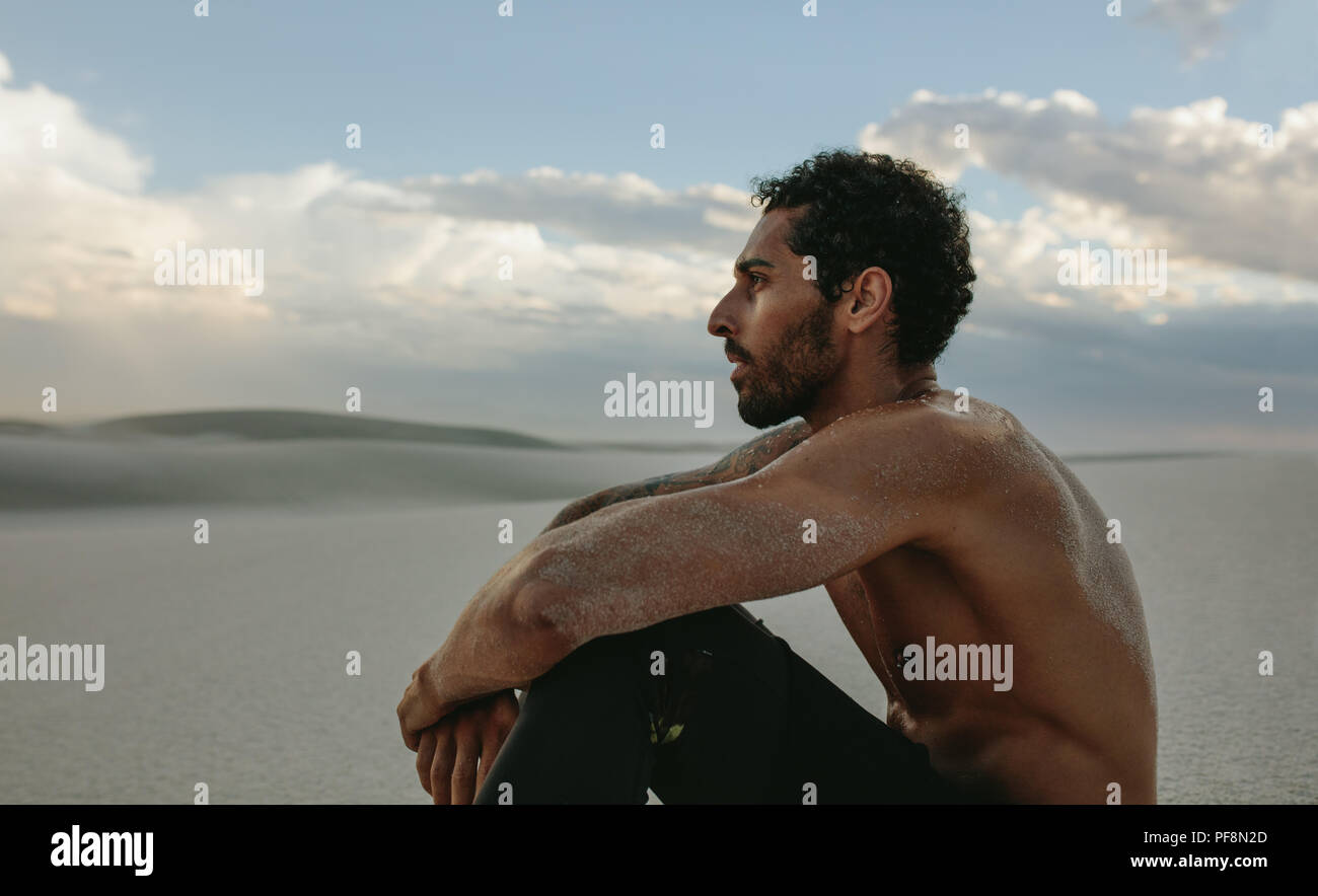 Side view of muscular male athlete sitting on desert sand and looking at a view. Tired man taking a break from training in desert. Stock Photo