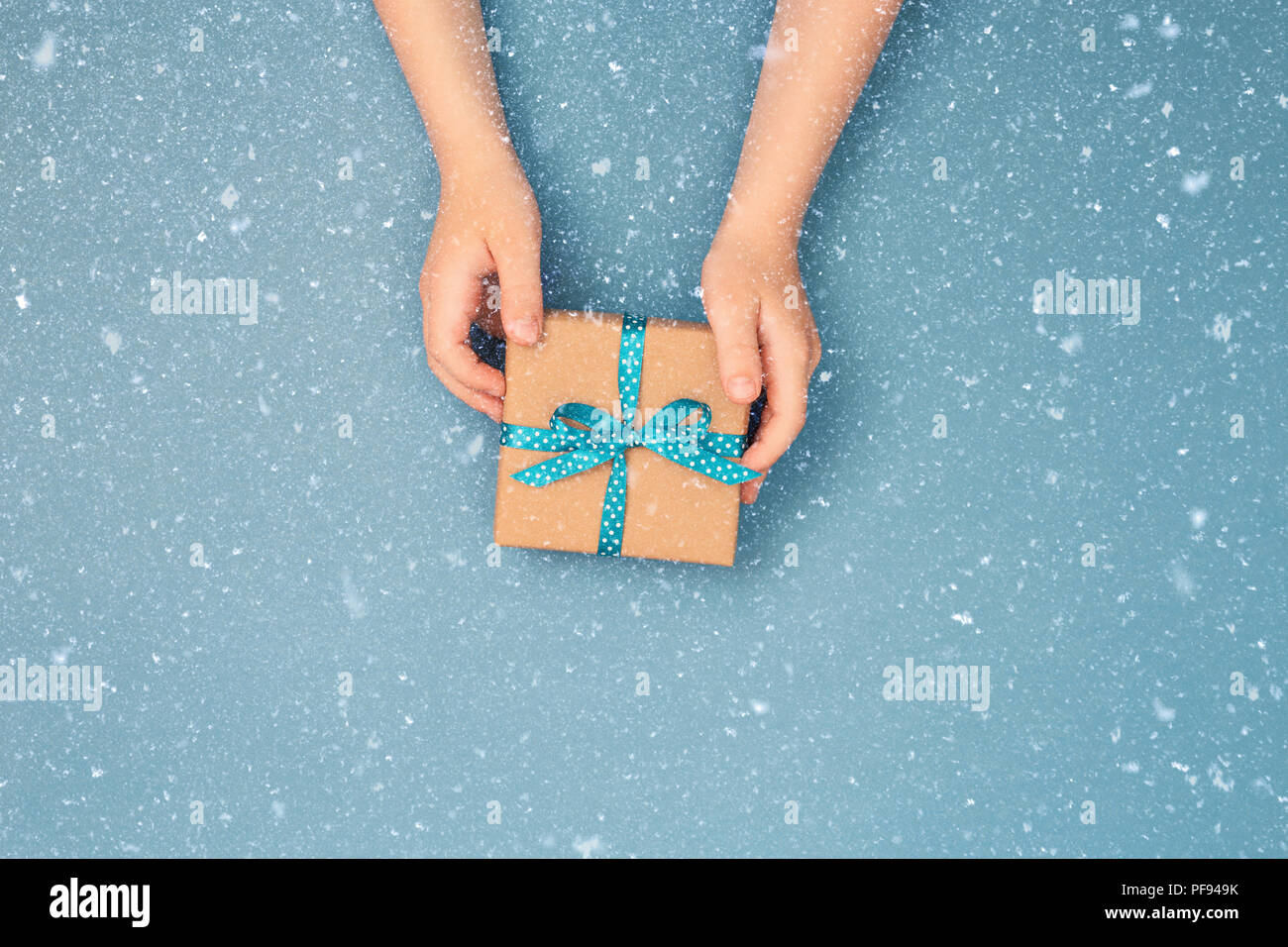 Child hands holding one gift box wrapped in kraft paper tied with blue ribbon in polka dots on blue grey background. Top view, place for text. Holiday concept. Stock Photo