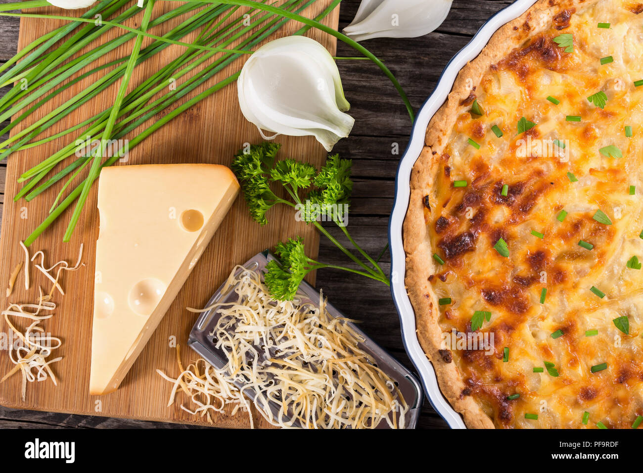 French onion cheese quiche or pie in a gratin dish sprinkled with parsley, with grater and ingredients on cutting board, authentic recipe, view from a Stock Photo