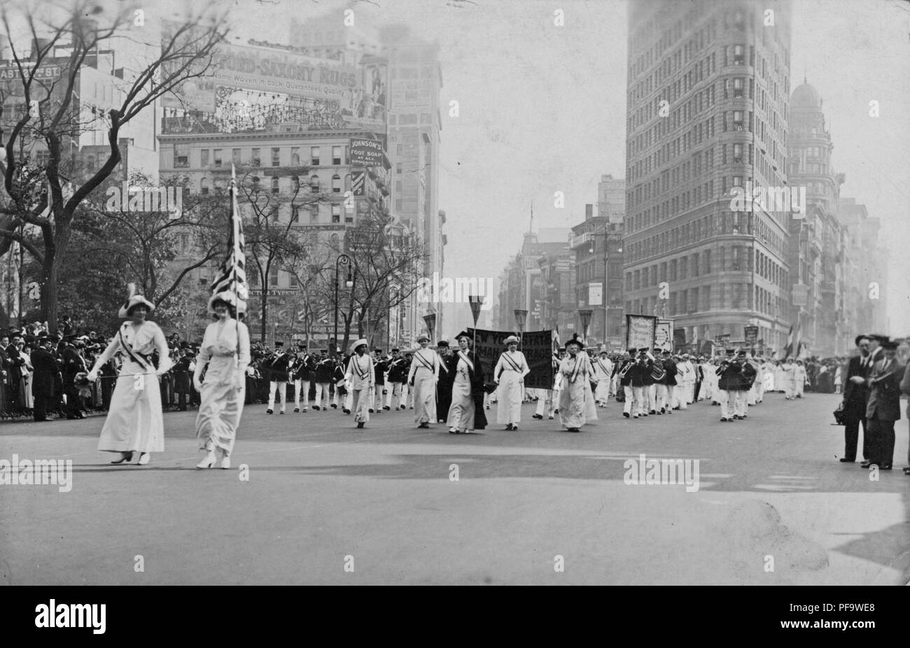 Black and white photograph showing several women, wearing white Edwardian clothing, and sashes, and holding a US flag and pennants, followed by a marching band, taking part in a suffrage parade in New York City, 1913. () Stock Photo