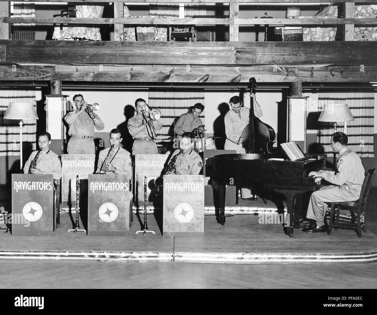 Black and white photograph, showing eight male members of the 'Navigators' band, wearing military clothes, sitting and standing on a low dais, with a wooden gallery overhead, and playing a variety of instruments, including horns, saxophone, bass, and piano, with a winged symbol visible on several musicians' sleeves suggesting an Air Force affiliation, likely photographed in Ohio, during the 1940s, 1945. () Stock Photo