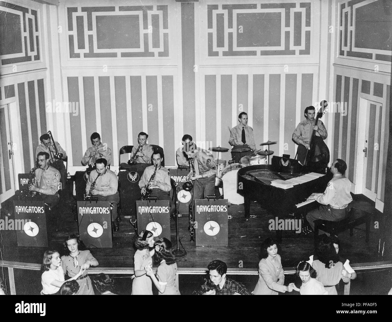 Black and white photograph, showing 11 male members of the 'Navigators' big band, wearing military clothes, sitting and standing on a dais, and playing a variety of instruments, including horns, saxophone, drums, bass, and piano, with a winged-star symbol on a stand at rear suggesting an Air Force affiliation, and with dancers, mostly female, visible in the foreground, likely photographed in Ohio, during World War II, 1945. () Stock Photo