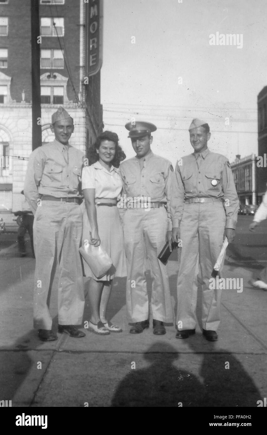 Black and white photograph showing a smiling young woman, wearing a skirt and holding a large clutch, standing between three smiling men dressed in military uniforms, all standing on a city street corner, in full-length view, photographed in Ohio, 1945. () Stock Photo