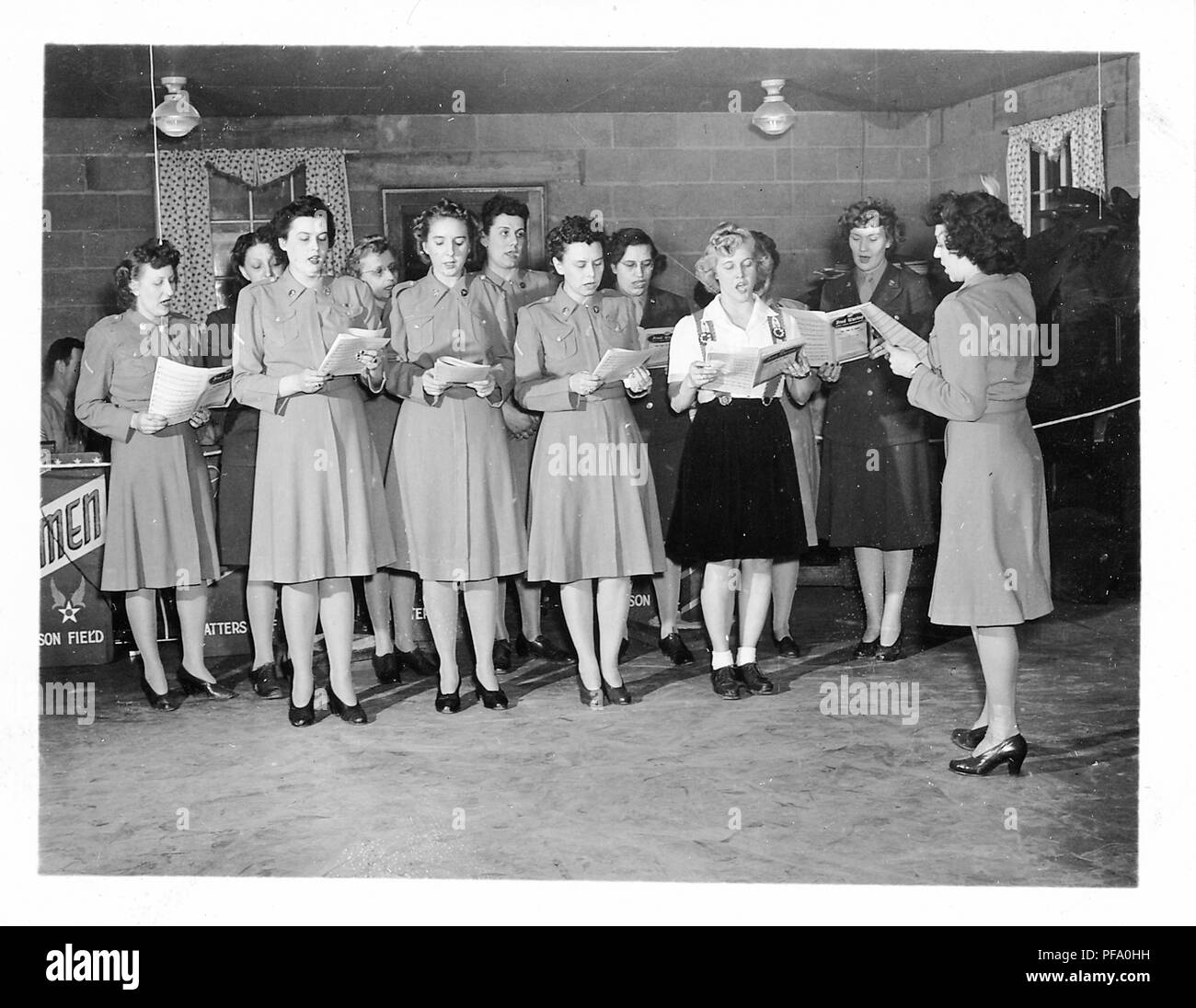 Black and white photograph, showing a group of women, most wearing military skirts and jackets, one in civilian skirt and top, standing indoors, and singing from musical scores that they hold in their hands, likely photographed in Ohio during World War II, 1945. () Stock Photo