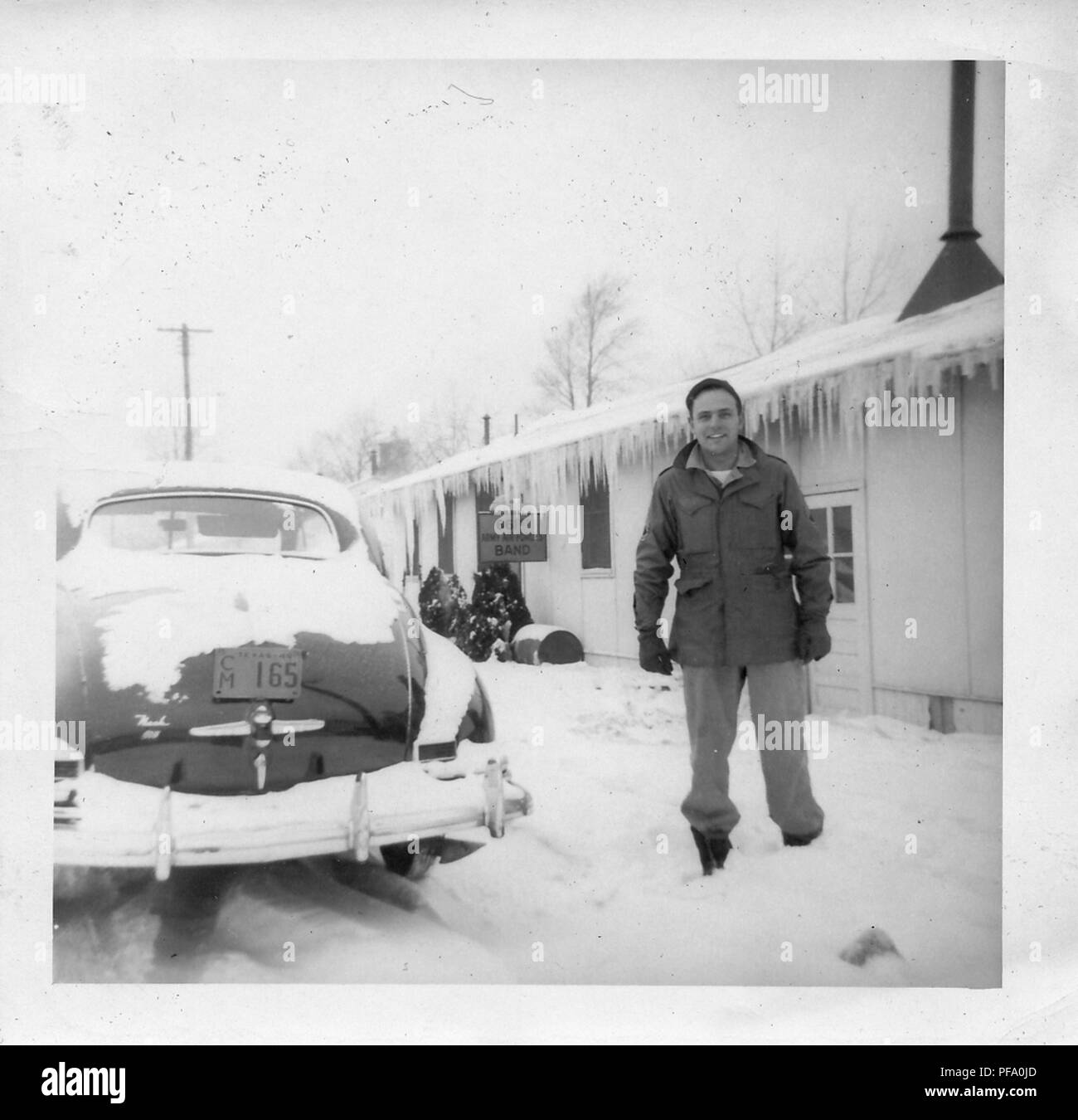 Black and white photograph, showing a smiling man, in full length, wearing military clothes and hat, standing outside in the snow, with a single story building in the background, a vintage car at left, and a sign on the building reading '661st Army Airforce Band, ' likely photographed in Ohio during World War II, 1945. () Stock Photo