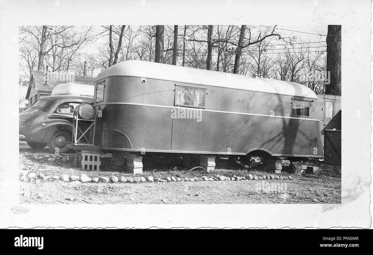 Black and white photograph, showing the side of a vintage caravan or towed trailer, resting on concrete blocks in a wintery landscape, with a Chevrolet sedan car, house, and more trailers in the background, likely photographed in Ohio in the decade following World War II, 1950. () Stock Photo