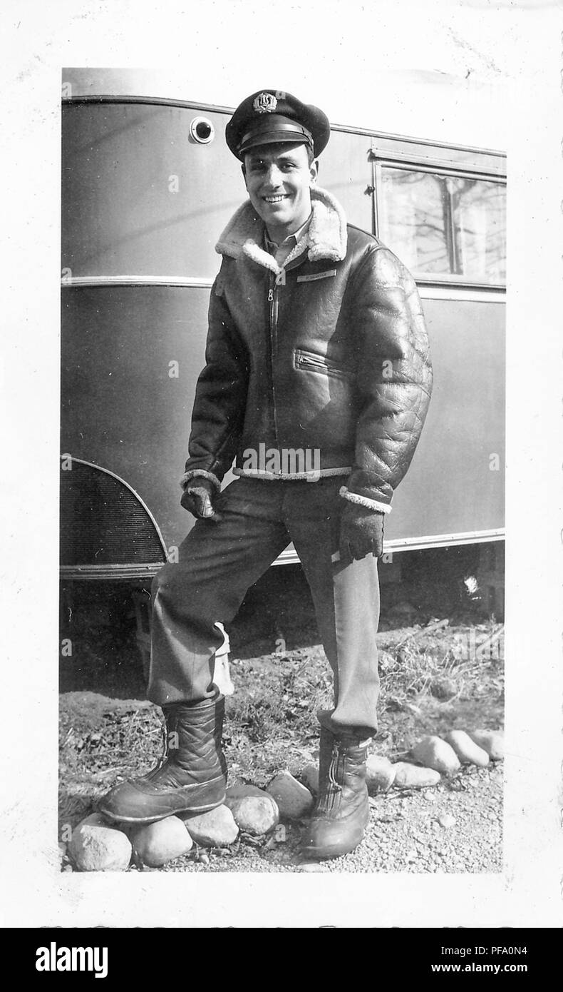 Black and white photograph, showing a smiling man, in full length, standing  outdoors, wearing an sheepskin lined, leather aviator or bomber jacket,  trousers, boots, gloves, and a cap with a military (presumably