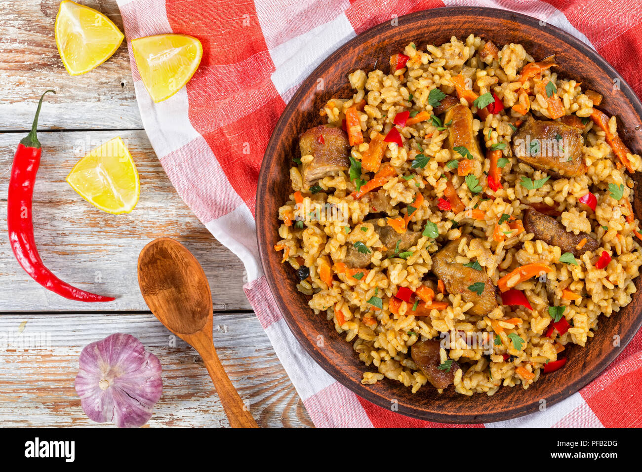 paella with meat, pepper, vegetables and spices on clay dish, kitchen towel,lemon slice, garlic, chili pepper and wooden spoon on white peeling paint Stock Photo