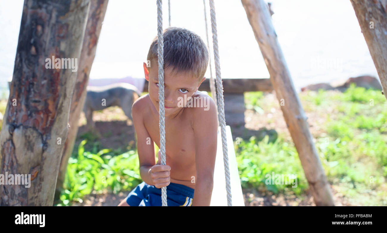 European boy sitting on a wooden old swing in a Park by the sea. Holidays in the village games in nature Stock Photo