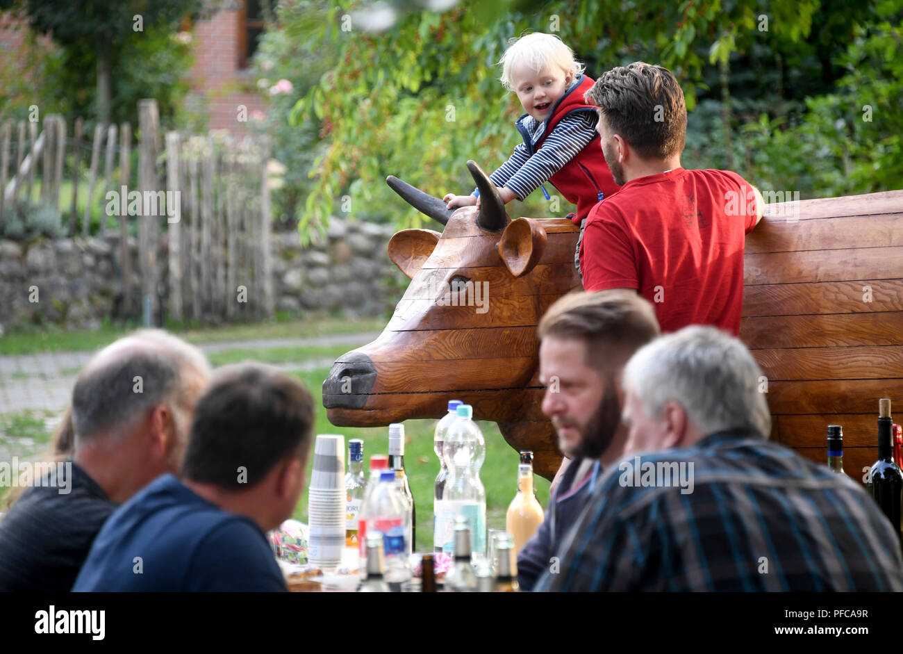 Aukrug-Homfeld, Germany. 19th Aug, 2018. The wooden cow 'Gerda Muh' stands behind visitors of a neighbourhood meeting. An anonymous artist created the wooden cow, since Whitsun it has appeared in changing gardens in the district of Homfeld. With this event, the initiator wants to stimulate the discussion about agriculture and the community feeling of the villagers. Credit: Carsten Rehder/dpa/Alamy Live News Stock Photo