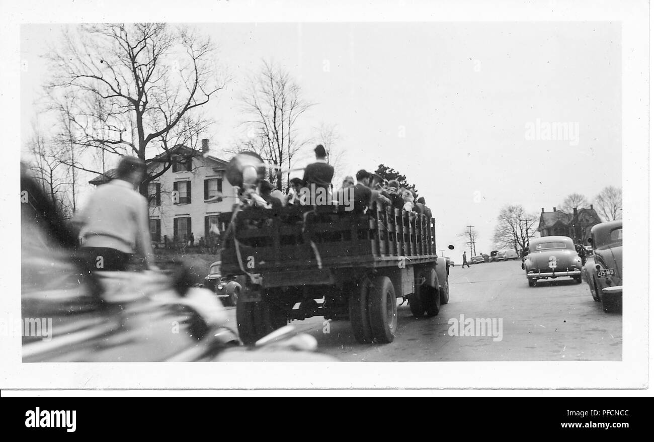 Black and white photograph, showing the rear of an open truck as it drives away, the truck bed is filled with members of a military band, some holding horns, with leafless trees, vintage cars, and houses visible on either side of the road, likely photographed in Ohio during celebrations marking the end of World War II, 1945. () Stock Photo