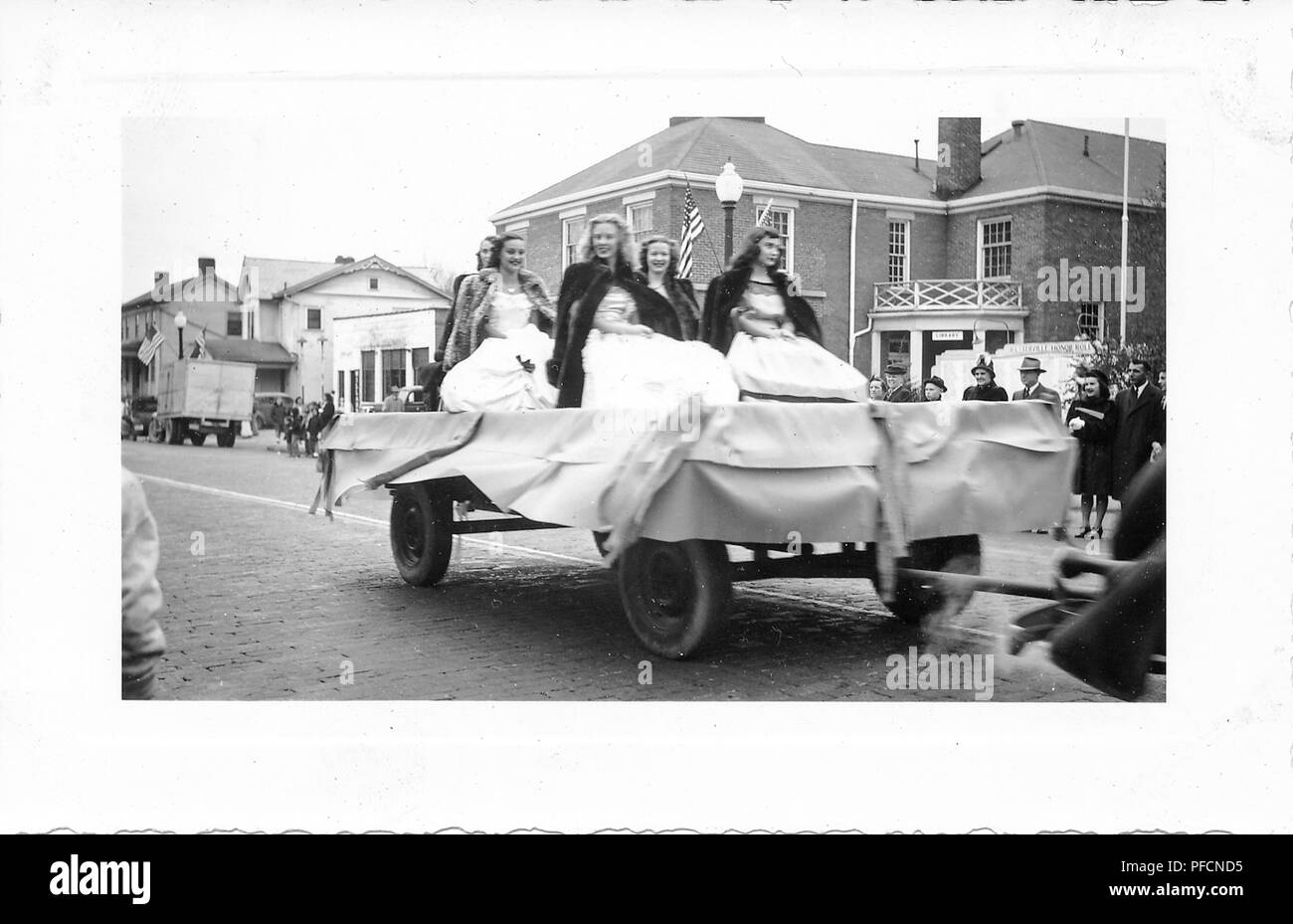 Black and white photograph, showing five girls, wearing ball gowns and fur coats, being pulled on an open parade float, with buildings, spectators, and American flags in the background, likely photographed in Ohio in the decade following World War II, 1945. () Stock Photo