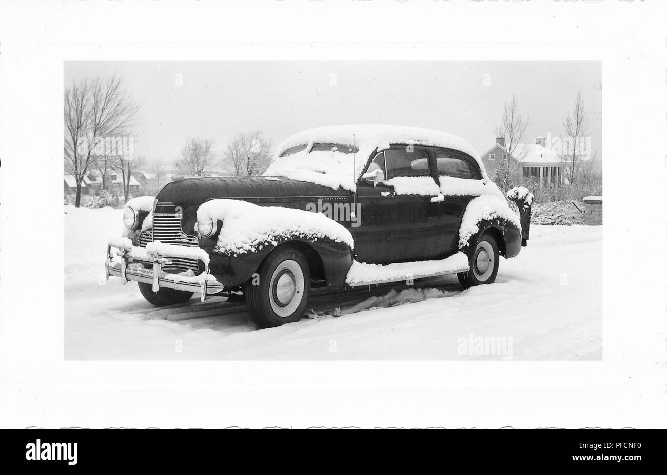 Black and white photograph, showing a three-quarter profile view of a dark, vintage, Chevrolet sedan car, parked outside in a wintery landscape, with freshly fallen snow on the roof, hubcaps, windshield, and running board, with leafless trees and houses in the background, likely photographed in Ohio in the decade following World War II, 1945. () Stock Photo