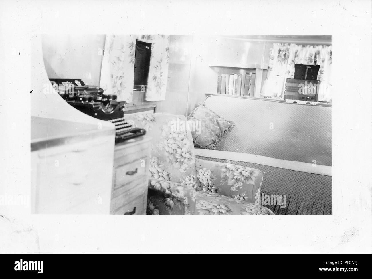 Black and white photograph, showing the cramped interior of a vintage caravan or trailer home, dominated by a black typewriter, floral easy chair, and chintz-covered sofa, with a small radio visible in the background, likely photographed in Ohio in the decade following World War II, 1950. () Stock Photo