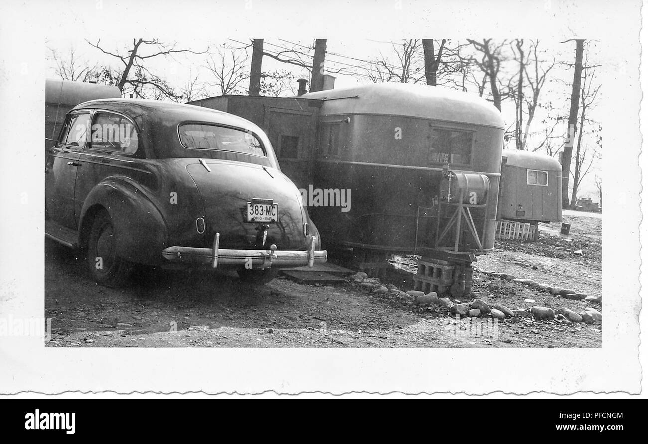 Black and white photograph, showing a vintage Chevrolet sedan car, with an Ohio license plated dated 1945, parked outside, in a wintery landscape, with a couple of caravans or towed trailers in the background, likely photographed in Ohio in the decade following World War II, 1950. () Stock Photo