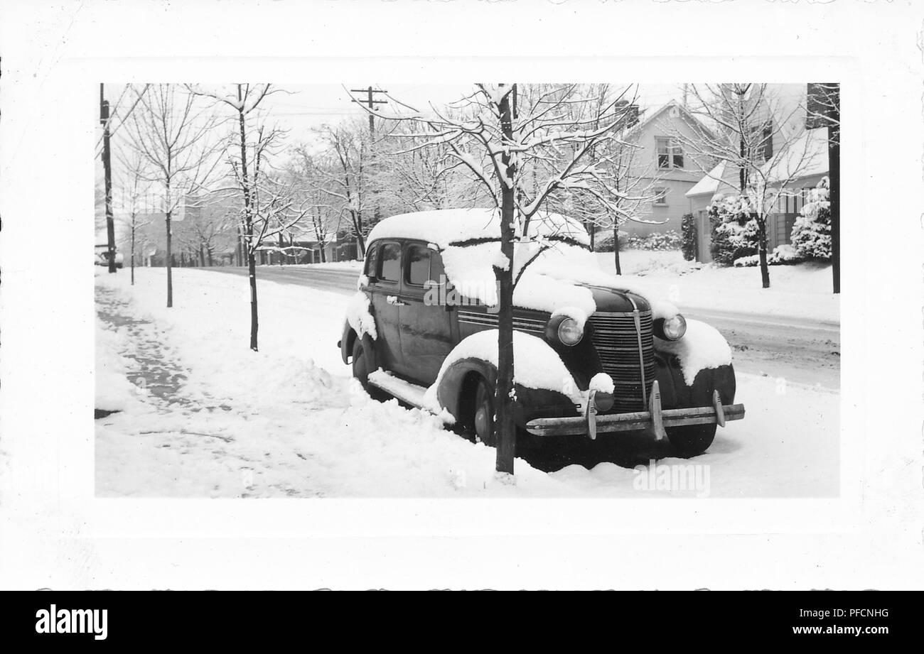 Black and white photograph, showing a slightly angled front view of a dark, vintage, Chevrolet sedan car, parked outside in a wintery landscape, with freshly fallen snow on the roof, hubcaps, windshield, and running board, with a leafless tree in the foreground and more trees and houses in the background, likely photographed in Ohio in the decade following World War II, 1945. () Stock Photo