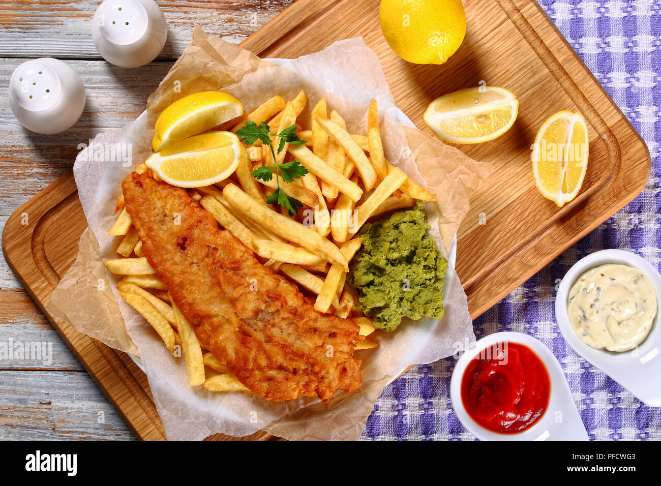 crispy fish and chips - fried cod, french fries, lemon slices, tartar sauce and mashed peas on plate on paper on old wooden table with rosemary in mor Stock Photo