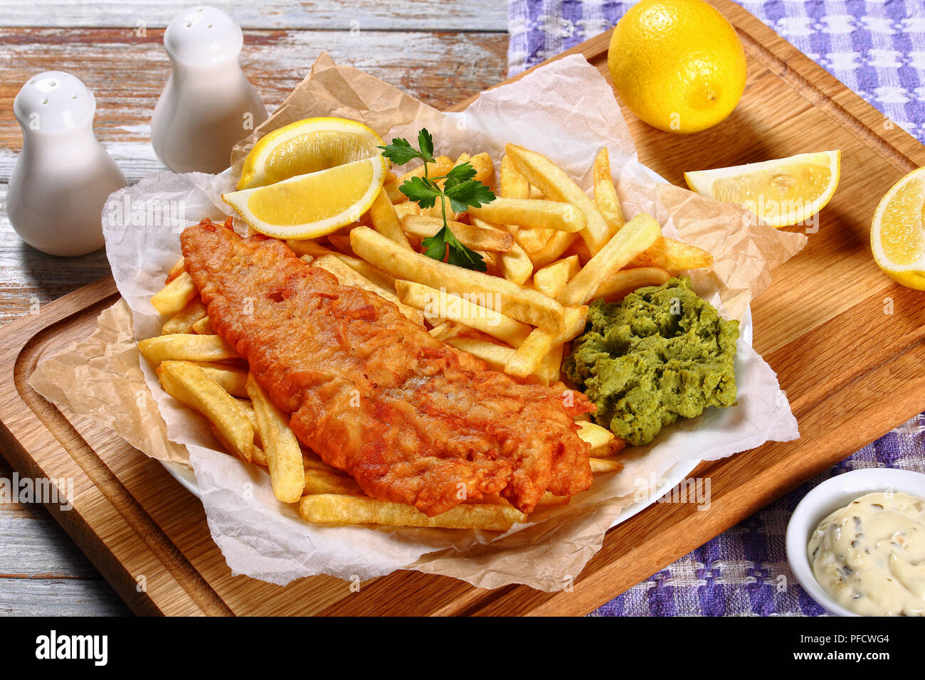 delicious crispy fish and chips - fried cod, french fries, lemon slices, tartar sauce and mushy peas on plate on paper. parsley and lemon wedges on wo Stock Photo