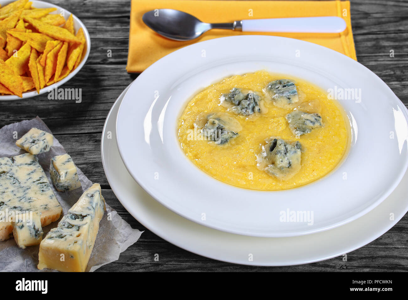 hot delicious creamy polenta with melted gorgonzola cheese on white plate with napkin and spoon. piece of gorgonzola on paper, saucer with cornmeal  s Stock Photo