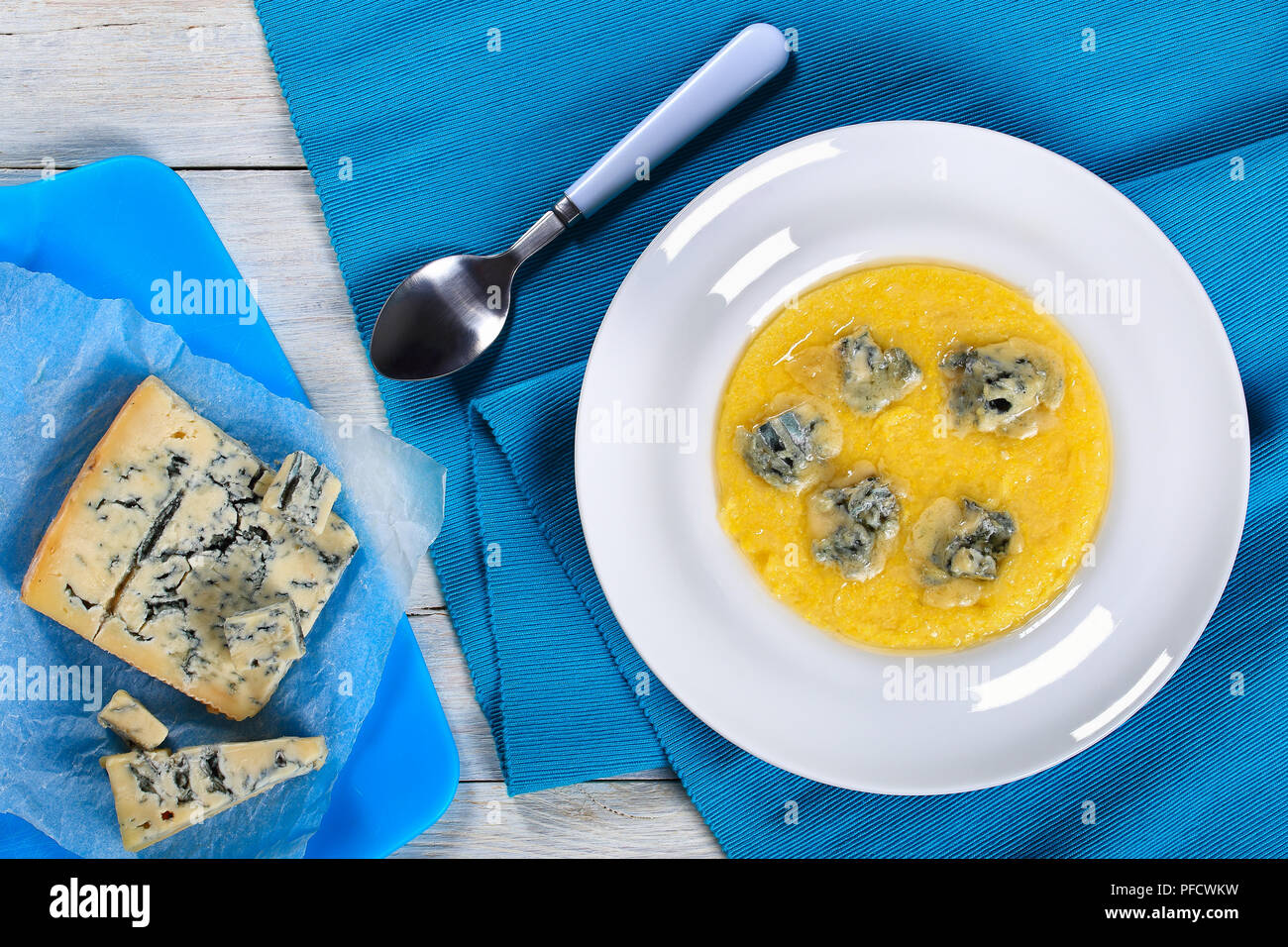 hot delicious creamy polenta with melted gorgonzola cheese on white plate on blue table mat with spoon. piece of gorgonzola  and chunks of cheese on c Stock Photo