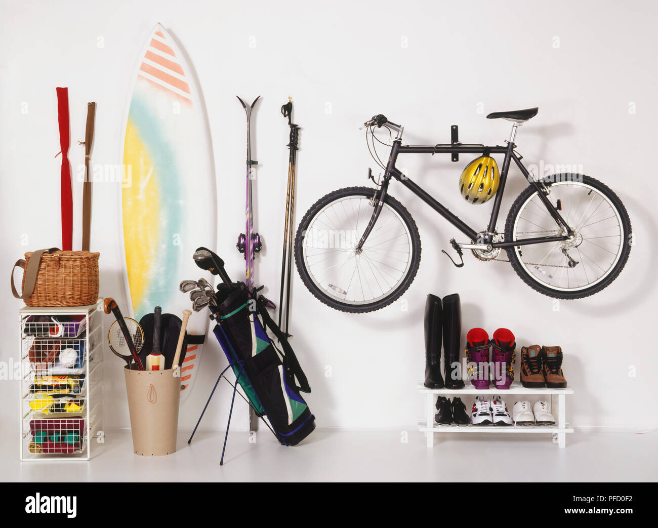 Bike fixed to wall, shoes stored on rack, skies and poles fixed on wall, golf clubs in caddy, surf board, hockey stick, front view. Stock Photo