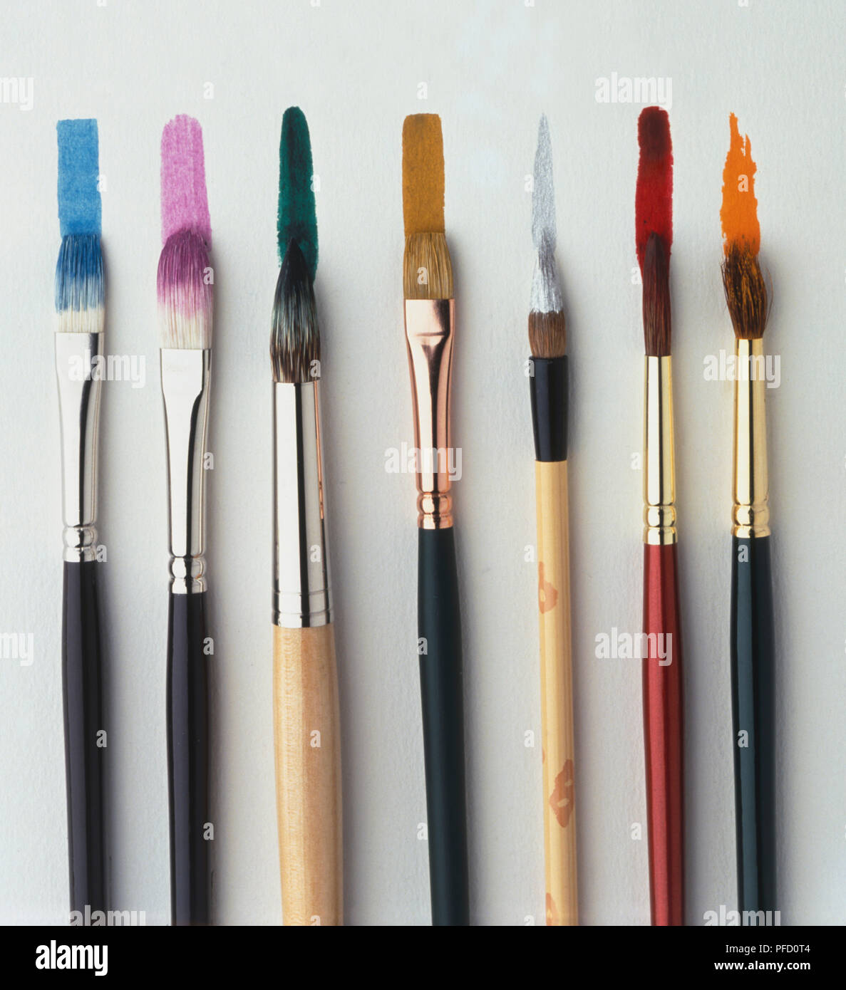 Seven paint brushes, different sizes, tips dipped in paint, strips of different colours painted on surface, selection of rectangular chips, providing colour samples. Stock Photo