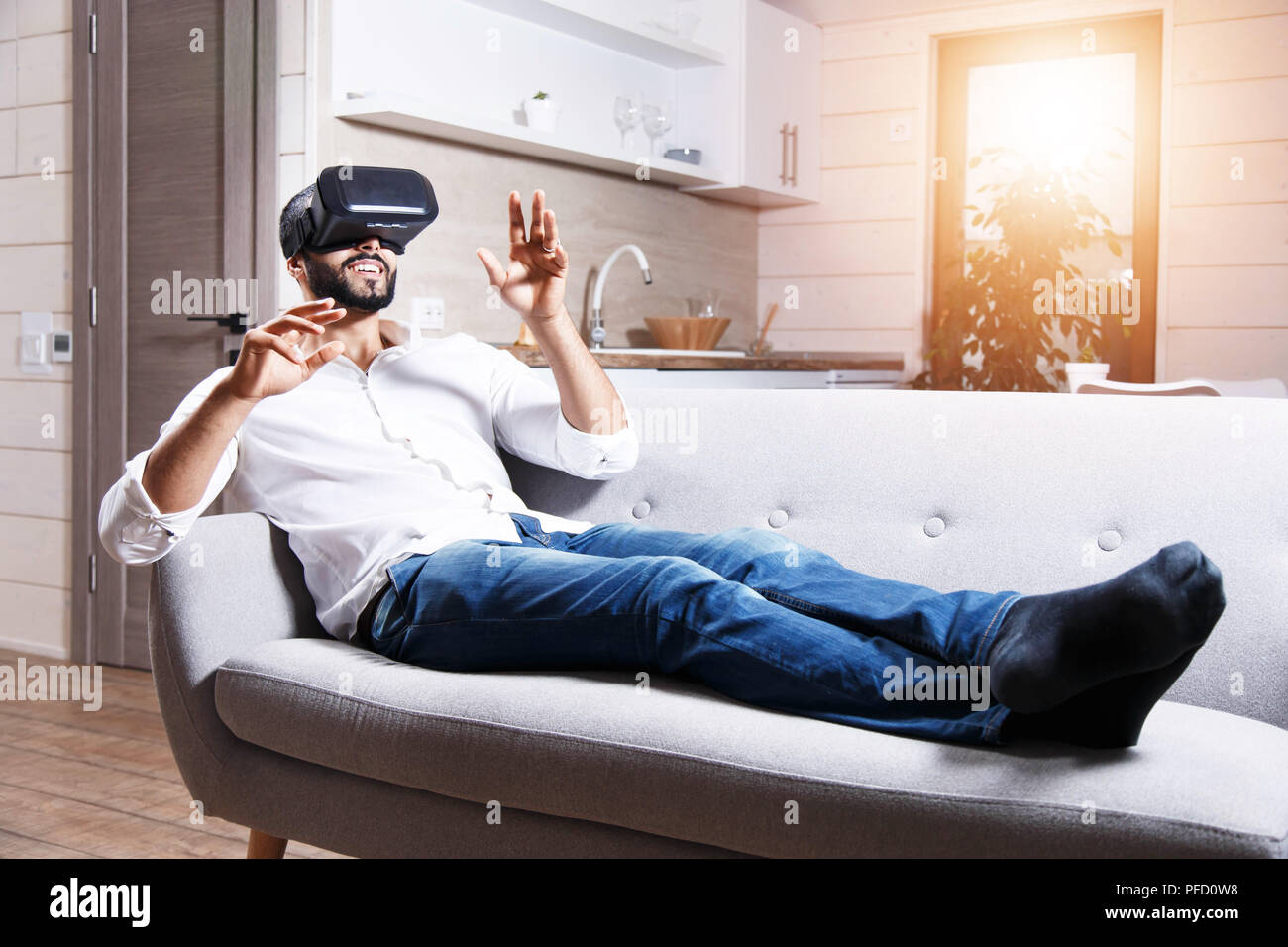 Multicultural bearded man laying on the sofa and using virtual reality goggles in the modern flat Stock Photo
