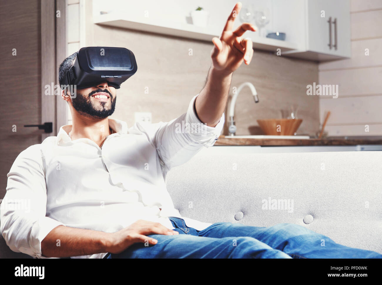 Multicultural bearded man laying on the sofa and using virtual reality goggles in the modern studio kitchen Stock Photo