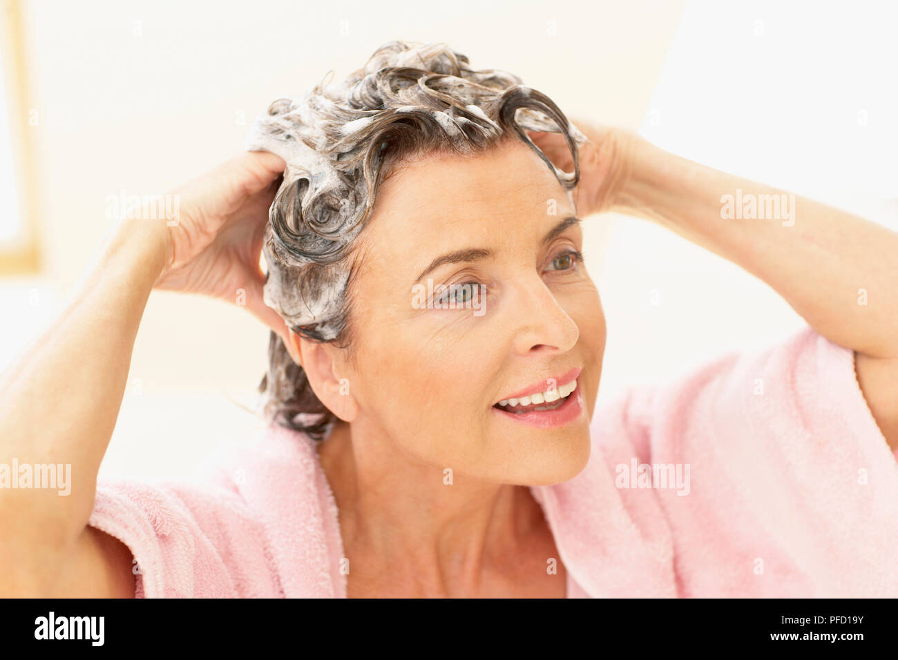 Woman smiling with eyes open, head and shoulders, in pink towelling robe, lathering hair with hands Stock Photo