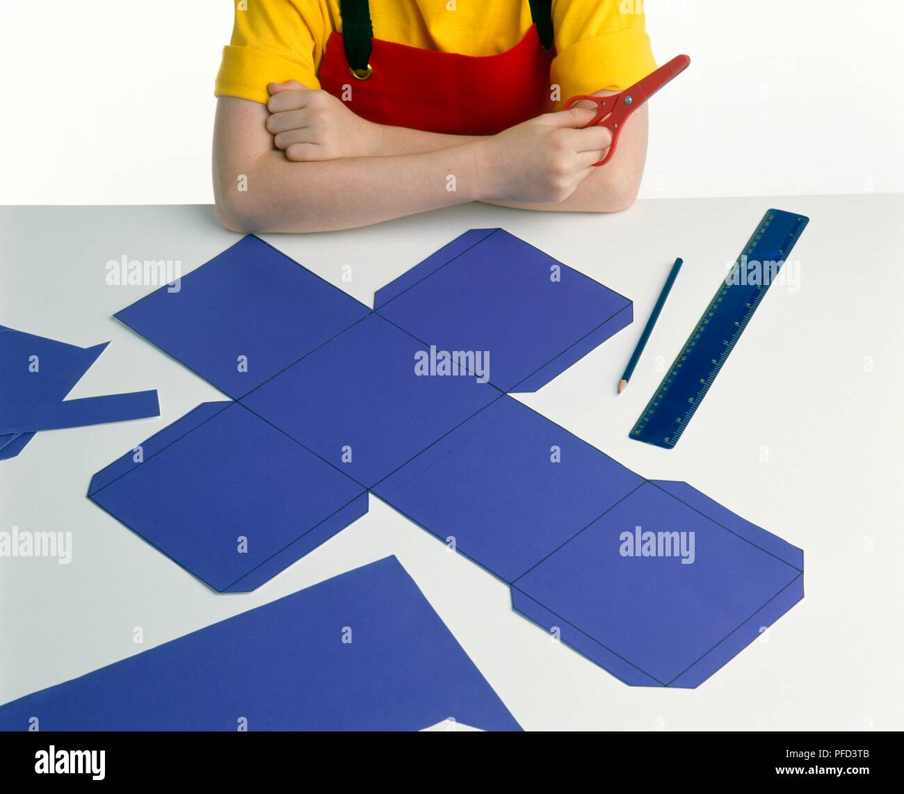 Making box out of card, joined squares of card with flaps on the side, ruler and pencil laid out on table, girl with her arms crossed holding pair of scissors, close-up Stock Photo