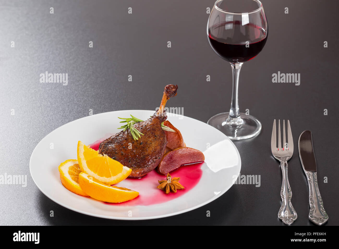 roasted duck leg in red wine with rosemary, orange and apple slices on white dish on black