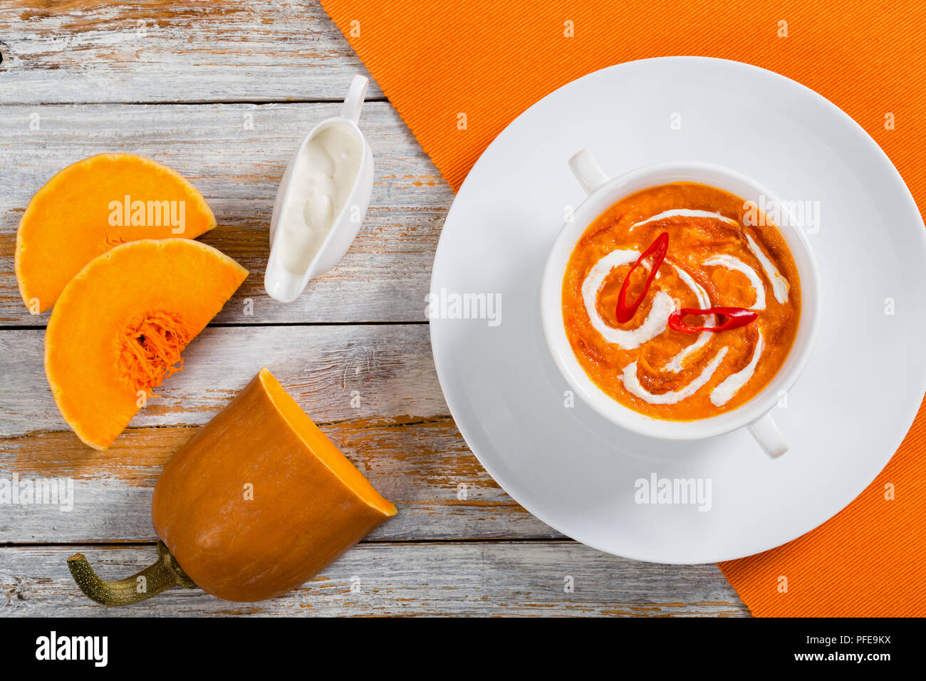 hot colorful Thai Pumpkin Soup puree with red thai paste, chili sauce and heavy cream in white cup on plate on table mat with cream in gravy boat and  Stock Photo