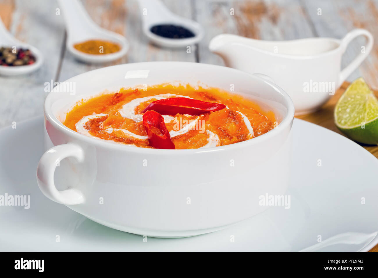 Thai Pumpkin Soup puree decorated with swirl in a little cream and chilli sauce in white cup on plate on chopping board with  lime slice and heavy cre Stock Photo
