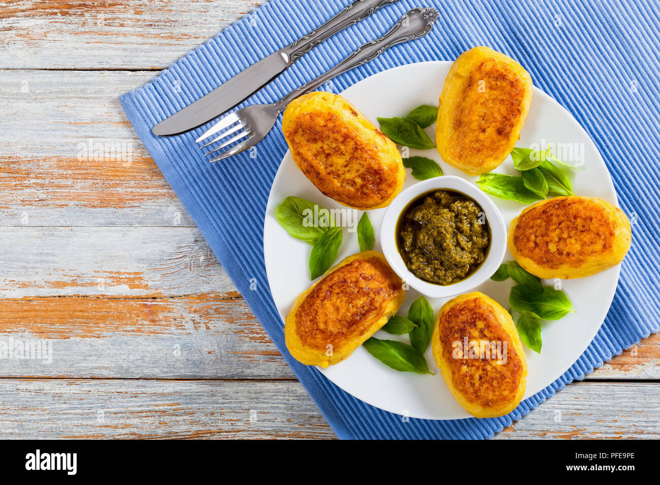 delicious Mashed Potato Pancakes with ground chicken Meat Filling on white dish with sauce pesto in gravy boat on table mat with fork and knife, view  Stock Photo